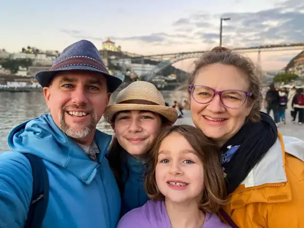 A family of four is posing for a selfie outdoors, with a scenic background of a river and a bridge, possibly during sunset. The father, on the left, is wearing a blue hoodie and a hat, while smiling at the camera. The mother, on the right, is wearing glasses, a yellow jacket, and a black scarf, also smiling. Between them are two children: the older one wearing a tan fedora and a darker jacket, and the younger one, in a light purple top, with long hair and a bright smile. The sky behind them is softly lit with warm colors, adding to the relaxed, happy atmosphere.