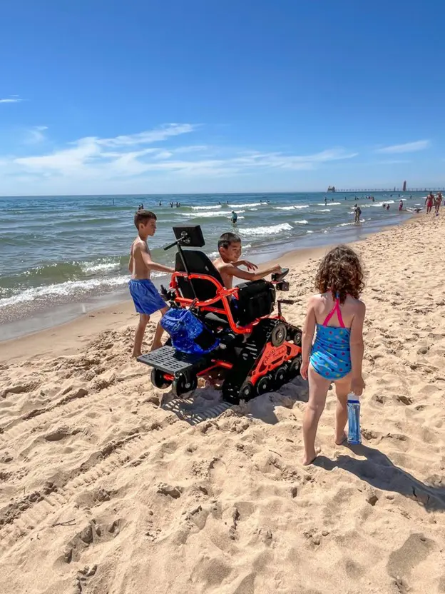 The image shows a bright, sunny beach with gentle waves rolling onto the sandy shore. In the foreground, a child is sitting in an orange all-terrain wheelchair with caterpillar-style tracks, designed for mobility on sand. Two other children are playing nearby, one standing in front of the wheelchair holding a water bottle, and the other is on the left side, closer to the water. The beach extends into the distance with several people wading and swimming in the ocean. The sky is clear and blue, with a pier visible in the background.