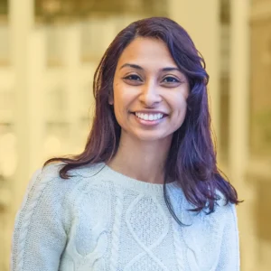 A woman with long, dark brown hair smiles warmly at the camera. She is wearing a light-colored, knitted sweater with a cable-knit pattern. The background is softly blurred, suggesting an indoor setting with natural light coming through large windows, creating a warm and inviting atmosphere.