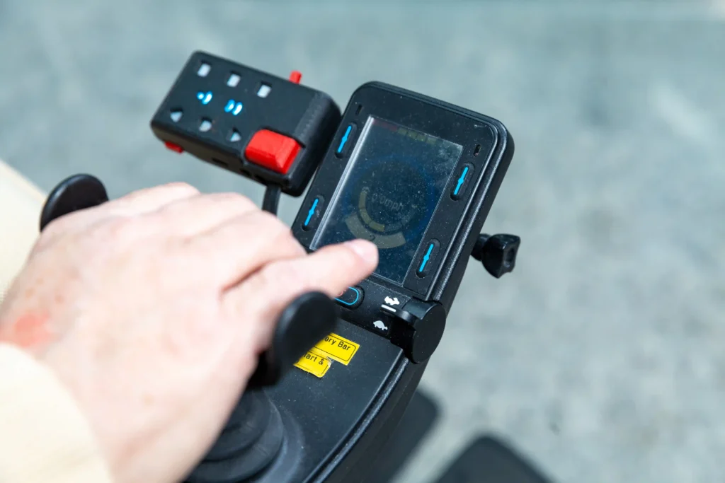 The image shows a close-up of a person's hand interacting with a control panel, likely on an electric mobility device such as a powered wheelchair or scooter. The control panel features a digital display, buttons, and a joystick. The display appears to show power or battery-related information. There are several blue-highlighted buttons surrounding the screen, along with a joystick to the left, and a prominent red switch at the top of the control module. The person's hand is resting on or near the joystick, indicating interaction with the controls.