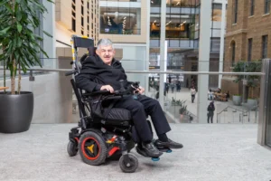 The image shows a man sitting in a motorized wheelchair in a modern indoor public space. The man, with gray hair and a mustache, is wearing a black coat and dark pants. He appears to be smiling while holding the controls of his wheelchair. The wheelchair has noticeable red accents on the wheels and footrests. The background features a large open atrium with glass panels, tall plants, and modern architecture with people walking or seated in the distance.