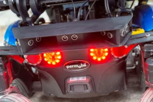 Close-up view of the rear section of a power wheelchair, featuring red brake lights and several sensors mounted above the lights. The wheelchair has a black frame with visible wires and components, and the Permobil logo is prominently displayed in the center.