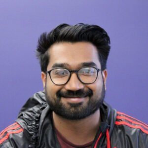 The image shows a smiling individual with short black hair and a beard. The person is wearing round glasses and a dark jacket with red accents. The background is a solid, muted purple color.
