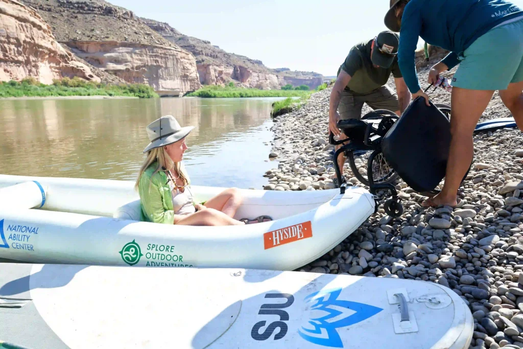 The image shows a woman sitting in an inflatable raft along the edge of a river with rocky shores. She is wearing a hat and a light green shirt, appearing relaxed as two people on the right side of the image handle a wheelchair. The background features scenic desert cliffs and a calm river, suggesting an outdoor adventure. The raft is branded with logos for "National Ability Center" and "Splore Outdoor Adventures," indicating that this might be part of an accessible outdoor program. The atmosphere is sunny and warm, with participants enjoying a day by the river.