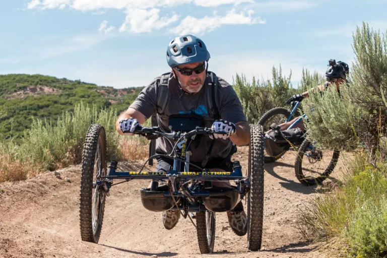 A person with paraplegia is riding an adaptive mountain bike on a dirt trail surrounded by greenery. They are wearing a helmet, sunglasses, gloves, and protective gear while navigating rugged terrain. Another rider on a conventional mountain bike follows closely behind. The scene takes place on a sunny day with a bright blue sky, offering a scenic outdoor adventure in a mountainous or hilly landscape.