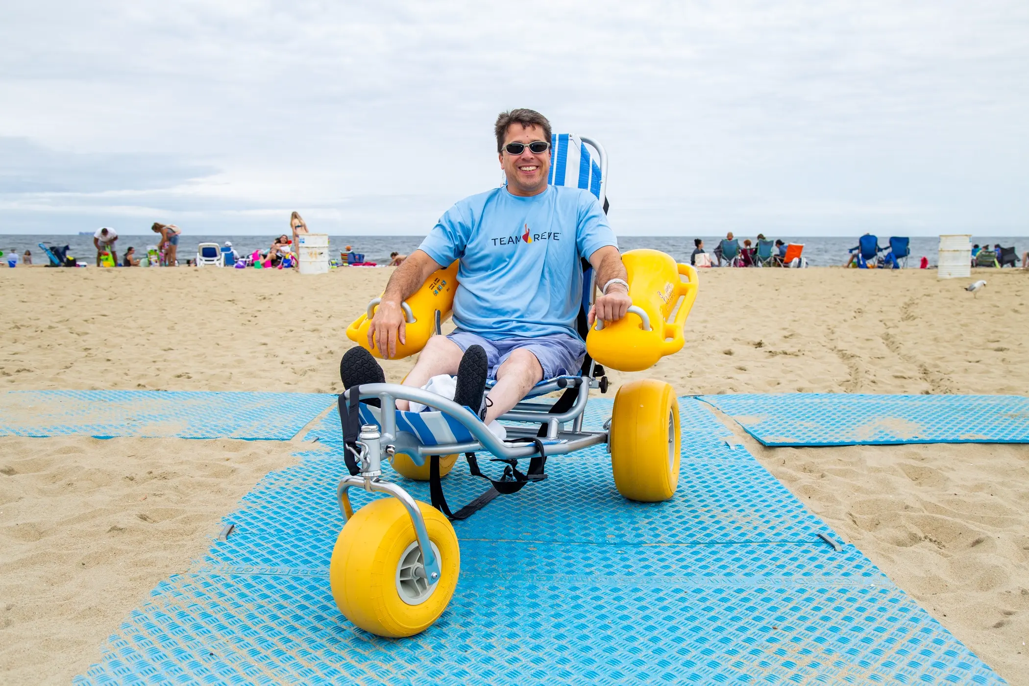 A man in a light blue "Team Reeve" t-shirt is sitting on a specialized beach wheelchair with large yellow balloon wheels. The beach in the background has a few people sitting in chairs and some walking along the shore. The wheelchair is positioned on blue matting that extends across the sand, allowing for accessibility. The man is smiling and wearing sunglasses, enjoying the day at the beach. The sky is cloudy, creating a soft light across the scene.