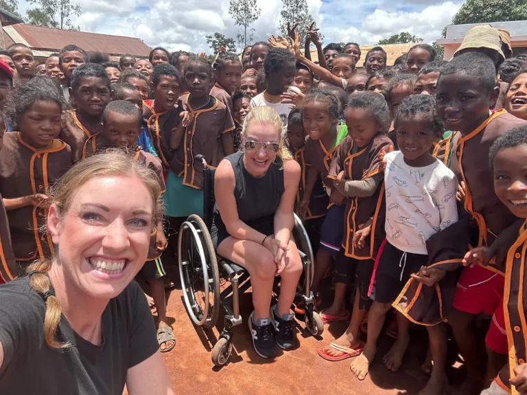 A joyful group photo featuring two women, one in a wheelchair, surrounded by a large group of smiling children. The woman in the foreground takes a selfie, beaming at the camera, while the other woman, seated in a wheelchair, is also smiling. The children around them are dressed in matching brown uniforms with orange accents and are closely gathered together, many smiling or waving. The setting appears to be outdoors on a sunny day, with a blue sky and some scattered clouds in the background. The children seem excited, standing in a schoolyard or similar location with rustic buildings behind them.