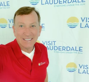 A man is smiling in front of a backdrop with the "Visit Lauderdale" logo, which also features the slogan "Everyone Under the Sun." He is wearing a red polo shirt with the same "Visit Lauderdale" logo embroidered on it. The backdrop and shirt emphasize a promotional theme for the Lauderdale area, likely related to tourism. The man has short, light brown hair and is looking directly at the camera.