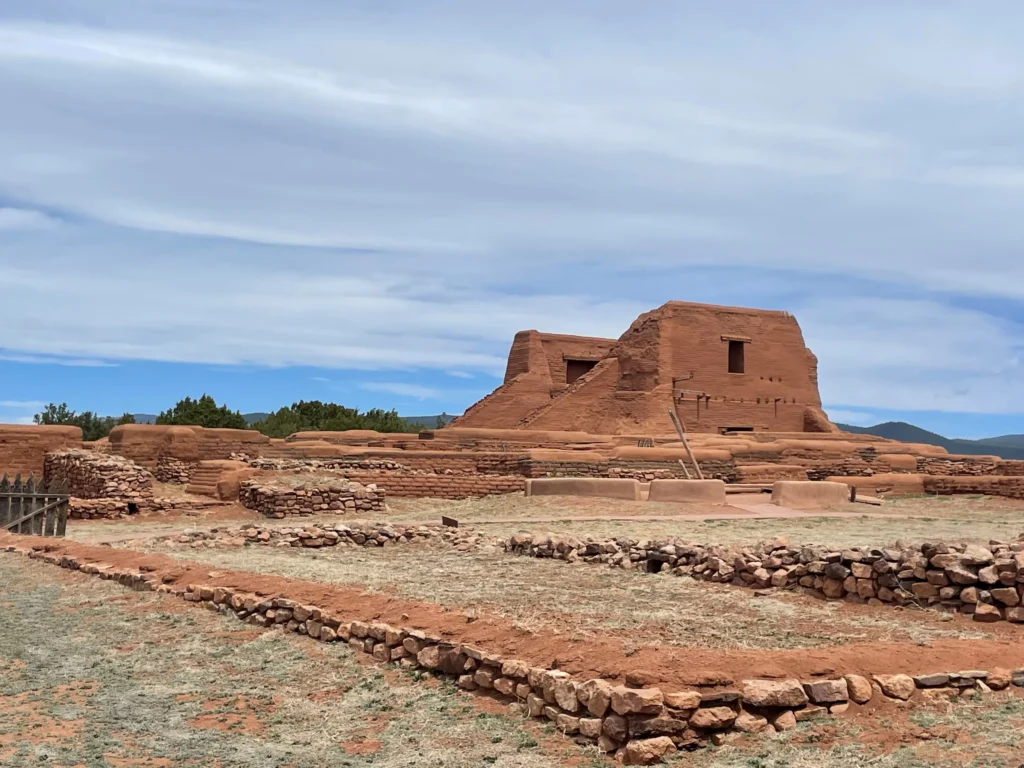 This image shows the ruins of an ancient adobe structure, likely in the American Southwest, surrounded by low stone walls and set against a backdrop of a lightly clouded sky. The building, constructed from reddish-brown adobe, appears weathered and eroded, though still retaining its characteristic flat-roofed, rectangular shape. The landscape is mostly arid, with dry grasses and some low bushes scattered around, giving the impression of a historic site, possibly a mission or pueblo ruins. The setting evokes a sense of history and connection to the indigenous or colonial past of the region.