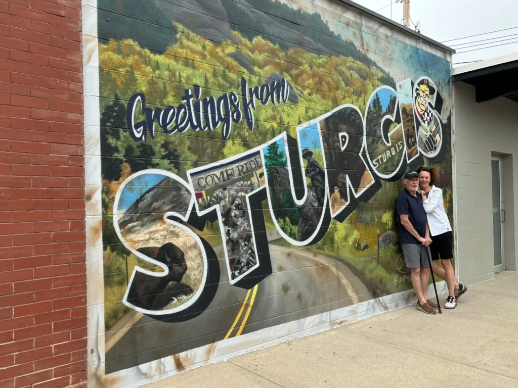 Two people standing in front of a colorful mural that reads 'Greetings from Sturgis.' The mural includes imagery of nature and a motorcyclist, paying homage to the town's well-known motorcycle culture. One person leans on a cane, while the other embraces them, smiling. They are positioned near the right side of the mural, next to a red brick wall on a city sidewalk.