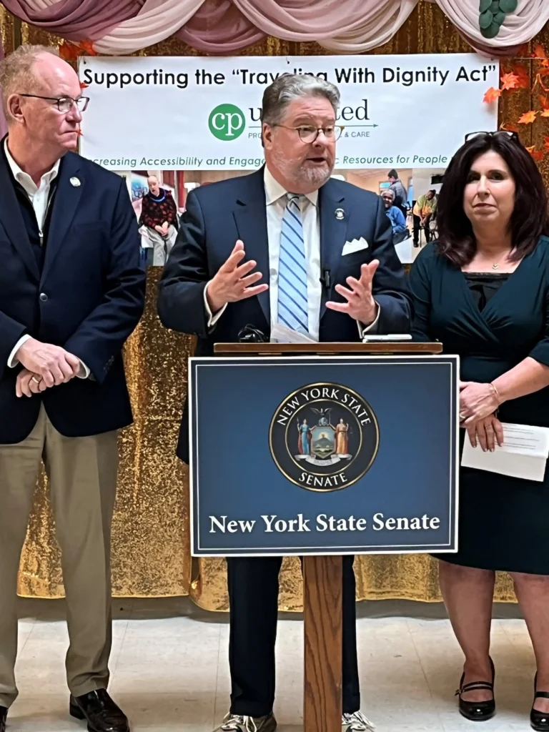 The image depicts three individuals standing behind a podium, which is labeled "New York State Senate." The person at the center is speaking and gesturing with both hands. Behind them is a banner supporting the "Traveling With Dignity Act," along with the logo of "CP Unlimited" (Cerebral Palsy Associations). The backdrop is adorned with decorative gold and pink drapes. The person speaking is flanked by a man on the left and a woman on the right, both of whom are standing attentively. All three individuals are dressed in formal business attire, suggesting that this is an official press conference or event related to the legislation.