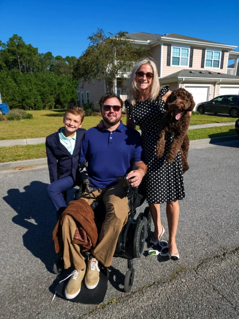A smiling family stands together outdoors on a sunny day. A man in a wheelchair is in the center, wearing sunglasses, a blue polo shirt, and khaki pants. A woman to his right, wearing a black and white polka dot dress, holds a brown dog in her arms. A young boy stands to the left of the man, wearing a blazer and smiling. They are standing on a residential street, with a house, trees, and blue skies in the background