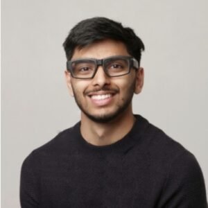 This is an image of a person smiling and wearing glasses. The person is dressed in a dark-colored sweater and has short, dark hair with a neat beard. The background is a plain light grey.