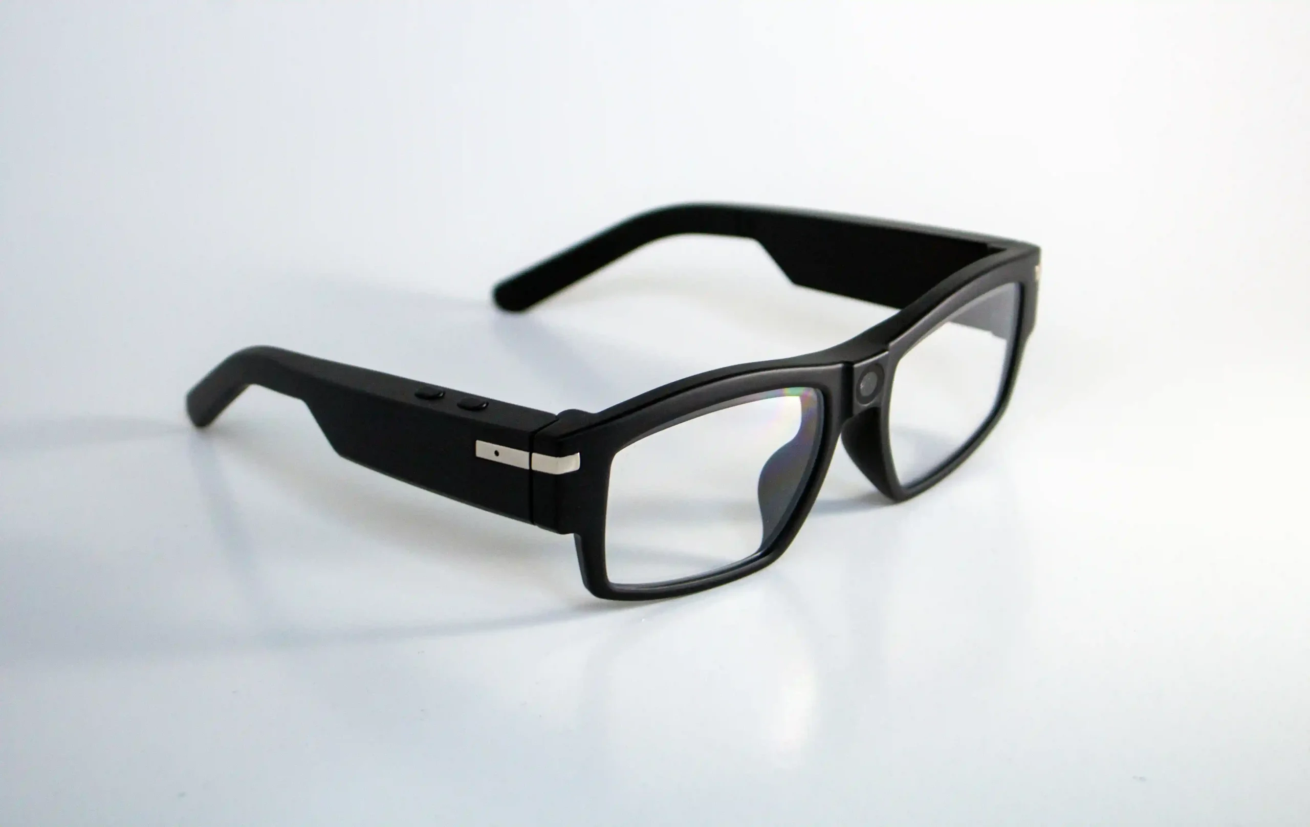 The image shows a pair of smart glasses with a black frame. The glasses have slightly thick arms, with two buttons visible on one side and a small metallic accent near the hinge. The lenses are clear, and a small circular sensor or camera is embedded at the top center of the frame, near the bridge of the glasses. The design appears sleek and modern, hinting at the presence of integrated technology.
