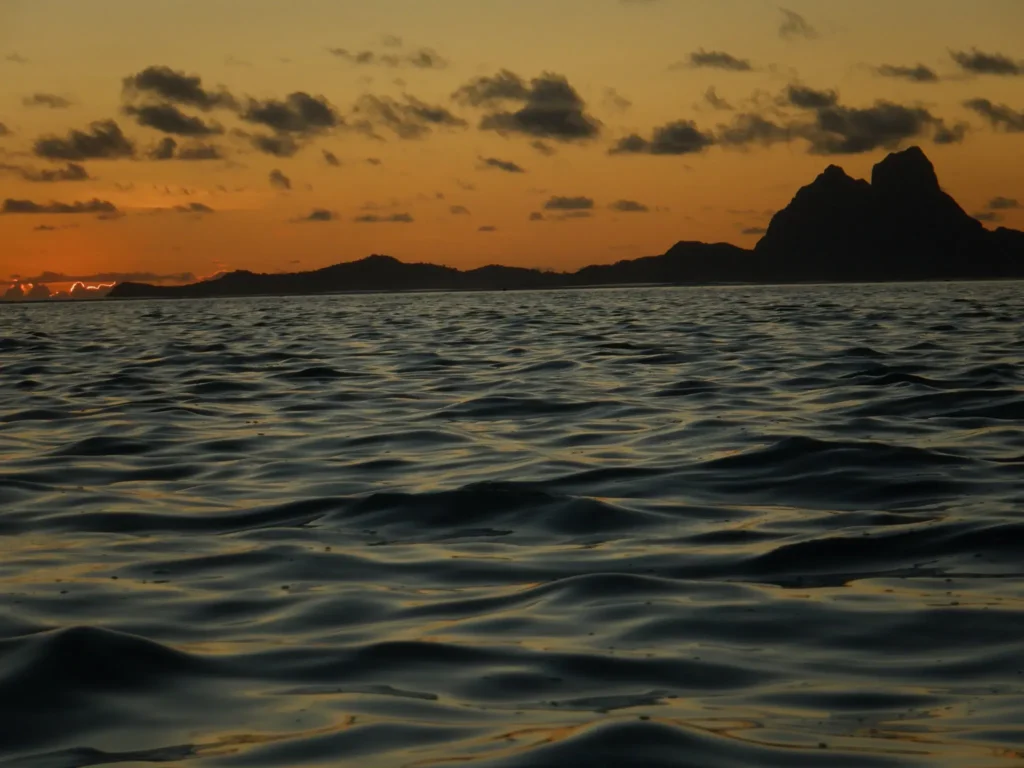 The image shows a serene ocean at sunset with gentle waves rippling across the surface. In the background, there's a silhouetted island with jagged peaks, and the sky is painted with hues of orange, red, and gold as the sun sets behind the clouds. The scene is peaceful, with a beautiful contrast between the darkened water and the warm colors of the sky.