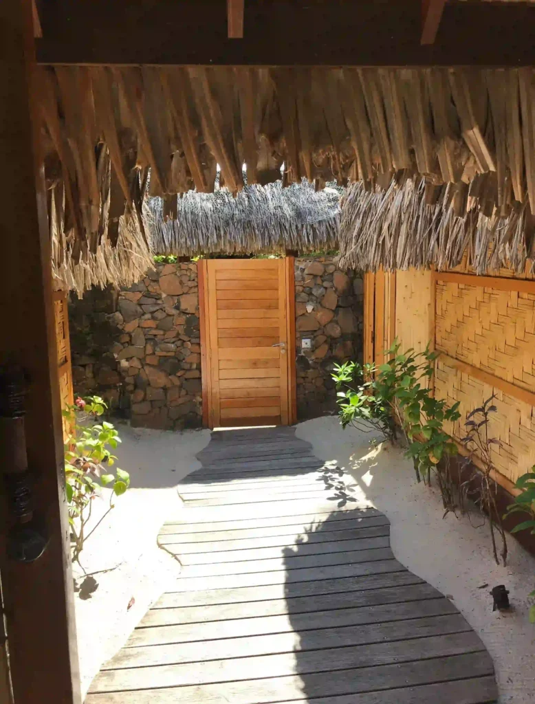 Tahaa Villa boardwalk: The image shows a wooden boardwalk path leading to a wooden door with horizontal panels. The path is set in an outdoor area with sand on either side, creating a tropical, beach-like vibe. The structure above and around the path features thatched roofing, with natural, dried leaves hanging down, giving it a rustic and natural appearance. The walls on either side are made from stone and bamboo-like materials, enhancing the tropical, island feel. Small plants and greenery line the sandy area beside the boardwalk.