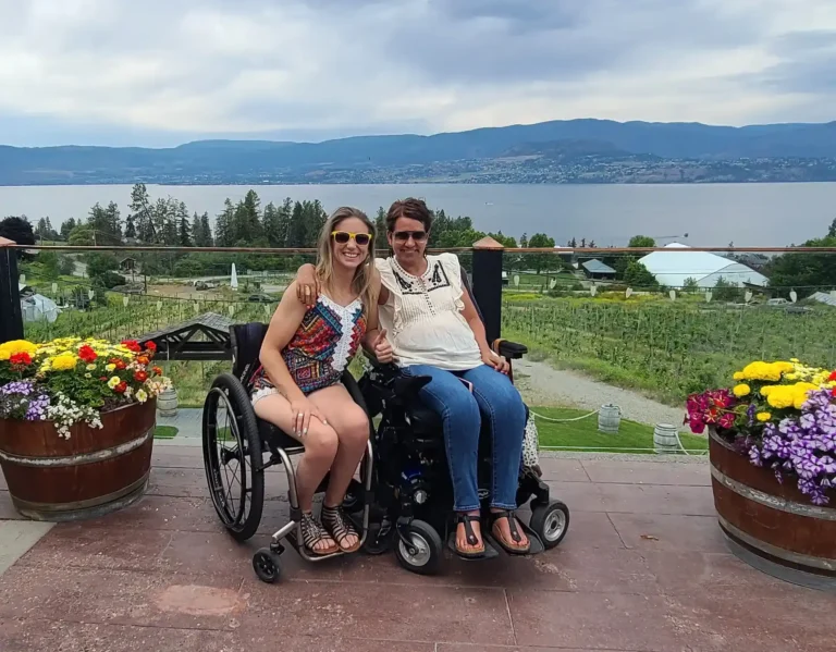 The image shows two women, both in wheelchairs, smiling and posing together in front of a scenic background. One woman, on the left, is wearing a colorful sleeveless outfit, sunglasses, and sandals, while the woman on the right is in a white top with jeans. They are sitting close, with their arms around each other, clearly enjoying their time. Behind them is a beautiful landscape featuring a vineyard, colorful flowers in wooden barrels, and a large lake with mountains in the distance under a slightly overcast sky.