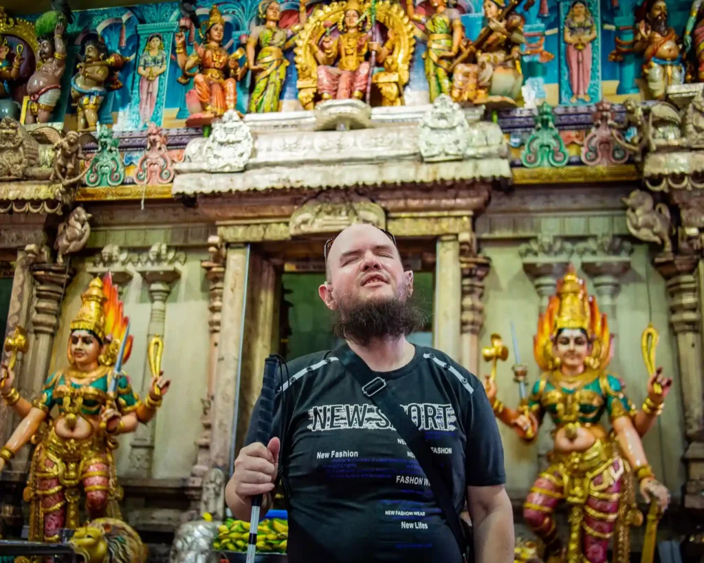 A man stands inside a Hindu temple, specifically in front of an ornate altar adorned with vibrant and detailed statues of deities. The statues depict Hindu gods and goddesses, intricately carved and painted with bright colors such as gold, green, and red. The man is wearing a black shirt that says "New Fashion" and has a beard. He appears to be visually impaired, holding a walking stick for guidance. His eyes are closed, and he seems to be experiencing the environment with a peaceful expression. The backdrop shows traditional temple architecture, with beautifully sculpted columns and vivid reliefs of deities.