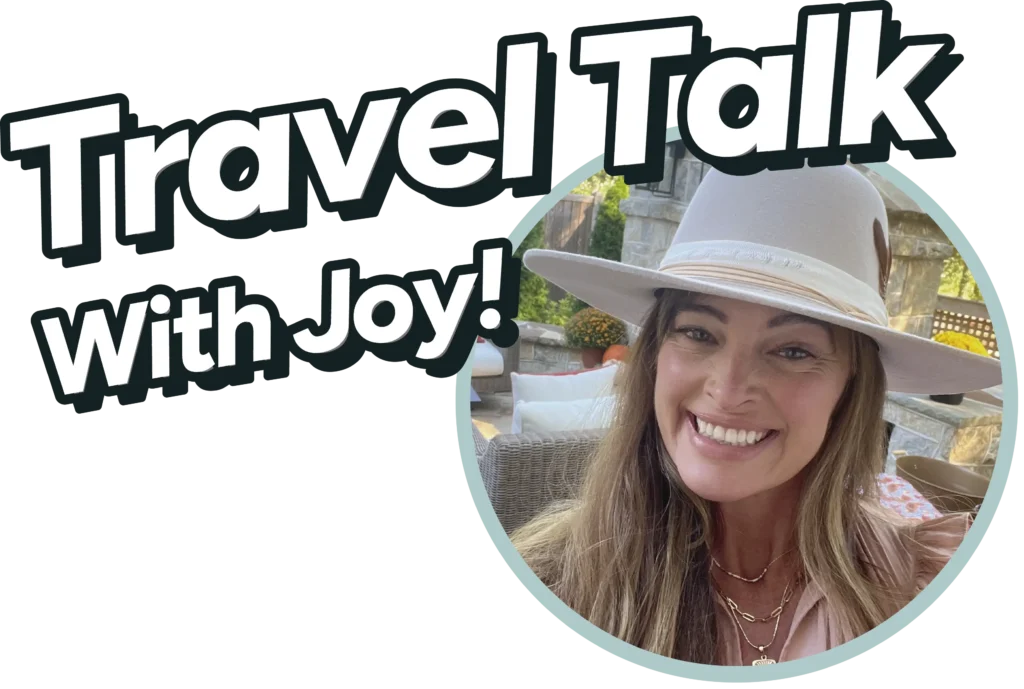 The image features a logo with the text "Travel Talk With Joy!" in a bold, fun font. To the right of the text, there is a circular photo of a smiling woman wearing a beige wide-brimmed hat. She has long hair, is sitting outdoors, and the background appears to be a cozy patio with a stone structure and some greenery. The overall vibe of the image is warm and inviting, suggesting a friendly and enjoyable conversation about travel.