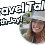 The image features a logo with the text "Travel Talk With Joy!" in a bold, fun font. To the right of the text, there is a circular photo of a smiling woman wearing a beige wide-brimmed hat. She has long hair, is sitting outdoors, and the background appears to be a cozy patio with a stone structure and some greenery. The overall vibe of the image is warm and inviting, suggesting a friendly and enjoyable conversation about travel.
