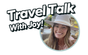 The image features a logo with the text "Travel Talk With Joy!" in a bold, fun font. To the right of the text, there is a circular photo of a smiling woman wearing a beige wide-brimmed hat. She has long hair, is sitting outdoors, and the background appears to be a cozy patio with a stone structure and some greenery. The overall vibe of the image is warm and inviting, suggesting a friendly and enjoyable conversation about travel.