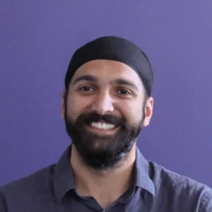A man is smiling in front of a solid purple background. He has a beard and is wearing a dark-colored turban along with a gray collared shirt. His expression appears cheerful and relaxed.