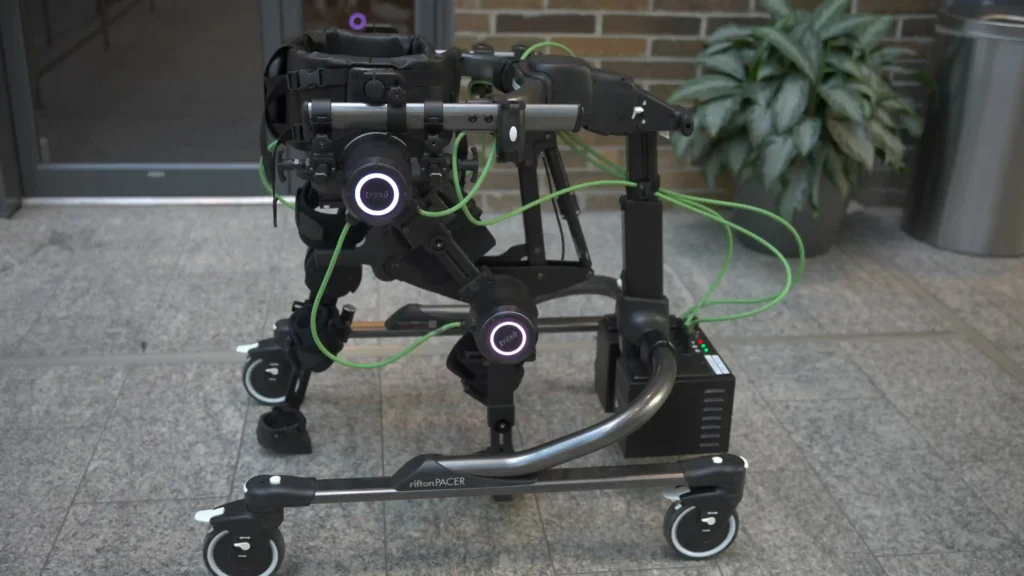 The image shows a robotic mobility aid device attached to a walker-like frame with four caster wheels. The device appears to be an exoskeleton designed to assist with walking or movement, featuring multiple mechanical joints and cables connecting various parts. The word "Trexo" is visible on the circular mechanisms around the robotic joints, which are illuminated by light rings. The frame supporting the exoskeleton is labeled "Rifton PACER" and includes components for support and balance. The device seems to be in an indoor setting with a tiled floor and plants in the background.
