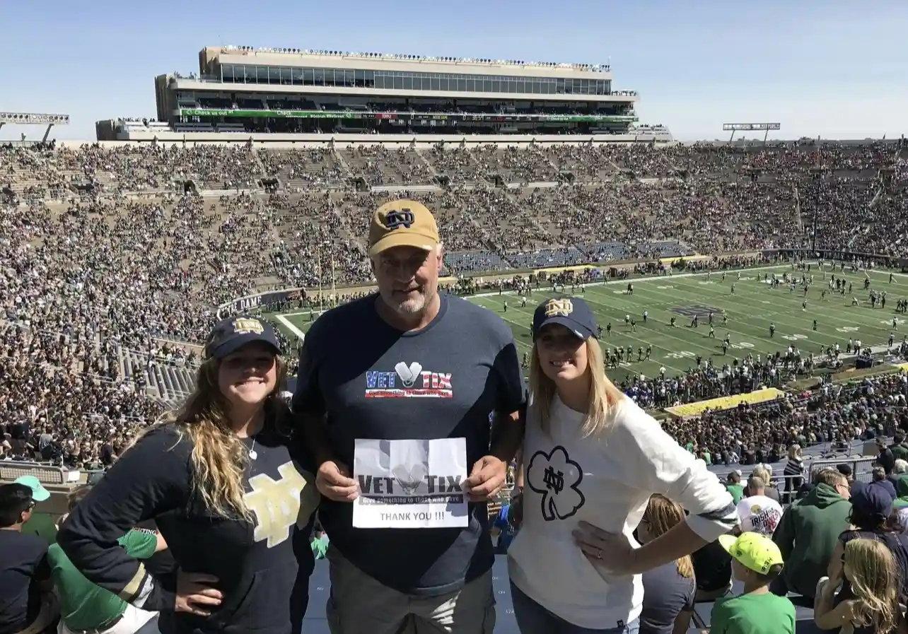 A group of three people is standing in a football stadium, with the field and crowd visible in the background. The person in the middle, wearing a navy blue Vet Tix shirt and a Notre Dame cap, is holding a sign that says 'Vet Tix' with a thank you message. The two people on either side, both wearing Notre Dame apparel, are smiling and posing with their hands on their hips. The stadium is filled with spectators, and the atmosphere is lively, indicating it is a game day.