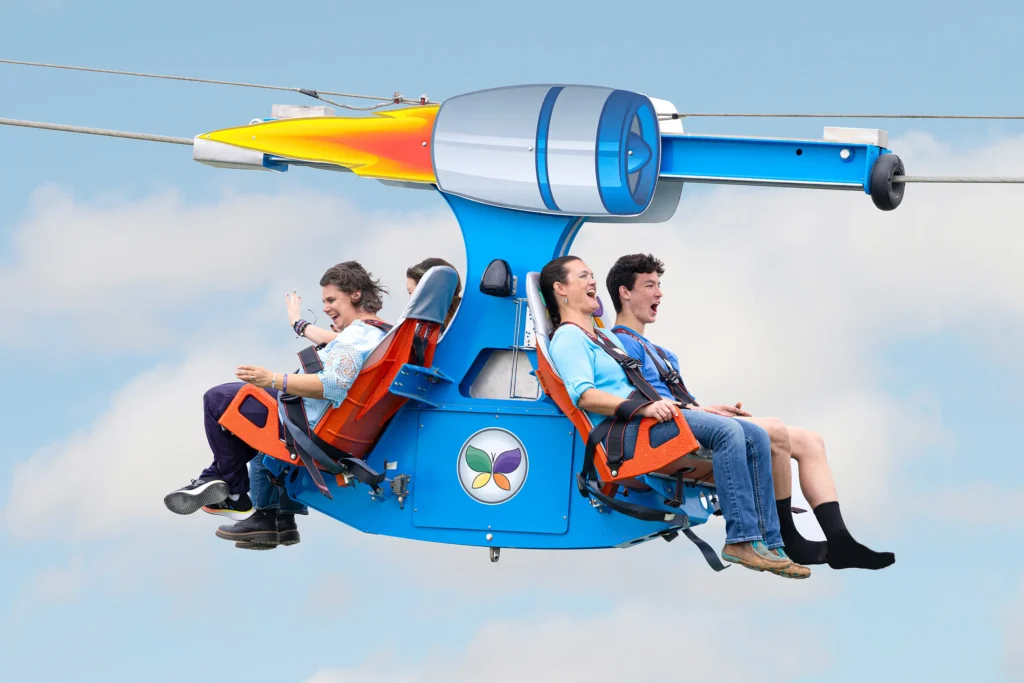 The image shows four people enjoying a ride on a futuristic zipline-style attraction. They are seated in two rows, each facing outward, and securely fastened with harnesses in bright orange seats. The ride appears to be suspended on cables with a sleek, blue and silver body resembling an aircraft engine or a small helicopter. The participants are visibly excited and smiling, with some raising their hands in the air. The background shows a bright, clear sky with scattered clouds, adding to the sense of height and thrill. A circular logo featuring four colored leaves is visible on the side of the ride.