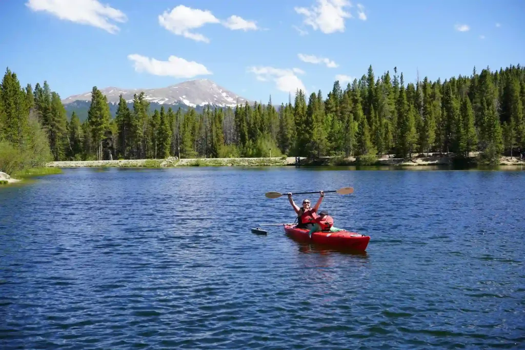 This image shows a joyful moment of two people kayaking on a calm lake surrounded by a lush forest and tall evergreen trees. The person in the front of the red kayak is raising their paddle high in celebration. In the background, a scenic mountain with patches of snow can be seen under a bright blue sky with a few white clouds. The overall atmosphere is peaceful and filled with natural beauty, with the individuals enjoying an outdoor adventure.