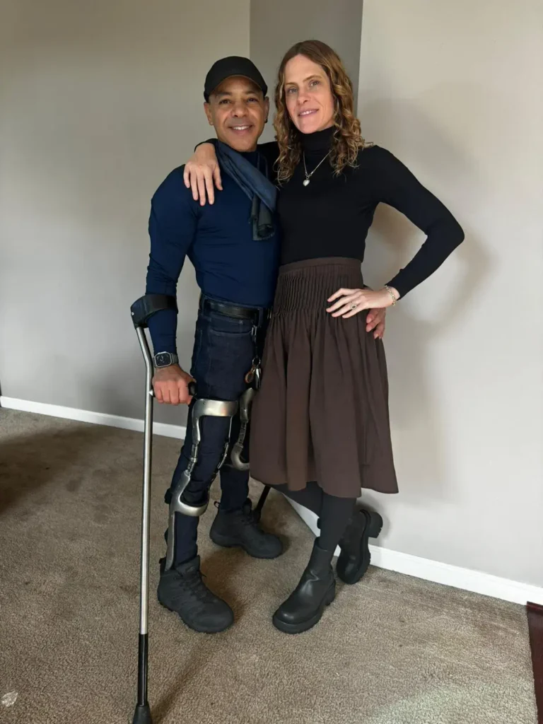 A man and a woman are standing together in a warmly lit room. The man is using a crutch and wearing supportive leg braces. He is smiling, dressed in a fitted dark blue top, a black cap, and black shoes. The woman has her arm around the man, wearing a black long-sleeved top, a brown skirt, black tights, and ankle boots. Both appear to be posing happily in a casual, indoor setting.