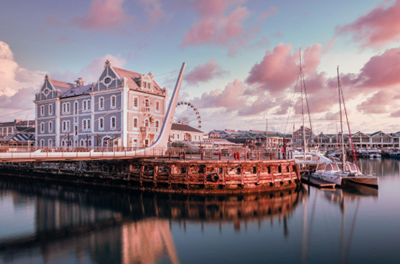 The image shows a serene waterfront scene during what appears to be sunrise or sunset, with a soft pink and purple sky reflected on calm waters. The focal point is an elegant historical building with white trim, likely a maritime or waterfront facility, alongside moored sailboats. In the background, a Ferris wheel is visible, suggesting a tourist or recreational area. The overall atmosphere is calm and picturesque, with pastel colors dominating the scene.