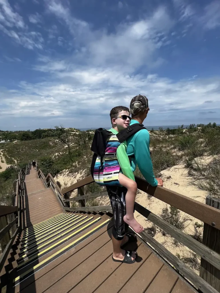 Two people are walking on a wooden boardwalk in a sandy, hilly landscape under a partly cloudy sky. The person in front (the father) is carrying the second person (their disabled son) on their back in a colorful backpack-like carrier. The person being carried is wearing sunglasses and smiling, while the carrier is wearing a cap and looking away. The boardwalk has steps that lead down a slope, with dunes and sparse vegetation in the background.