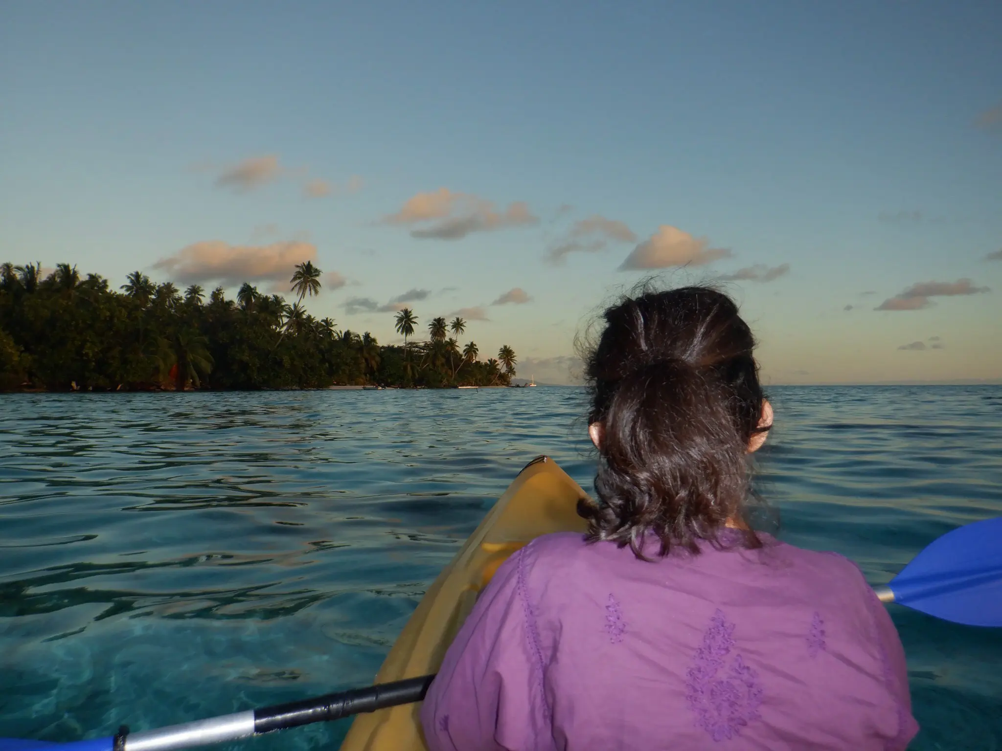 A woman is kayaking in calm waters during sunset or early evening. She is seated in a yellow kayak with a blue paddle, wearing a purple top, and her hair is tied in a ponytail. The view shows her from behind as she heads towards a lush tropical island with tall palm trees. The sky is clear with a few clouds, and the water reflects the soft colors of the sky, creating a serene and peaceful atmosphere.