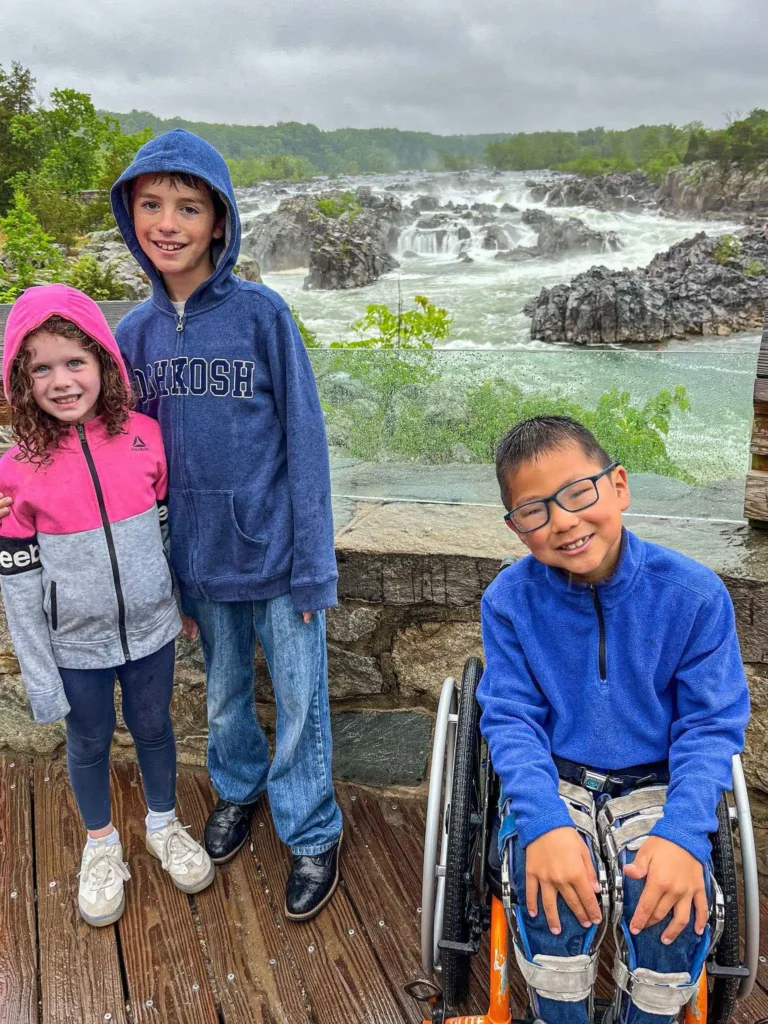 Three children posing together outdoors, with a scenic view of cascading waterfalls in the background. The boy on the left wears a blue hoodie and jeans, standing next to a younger girl dressed in a pink hoodie and leggings. On the right, a boy in a blue jacket sits smiling in a wheelchair. The weather appears misty or rainy, as the wooden boardwalk beneath them looks damp, and the children are wearing hooded jackets