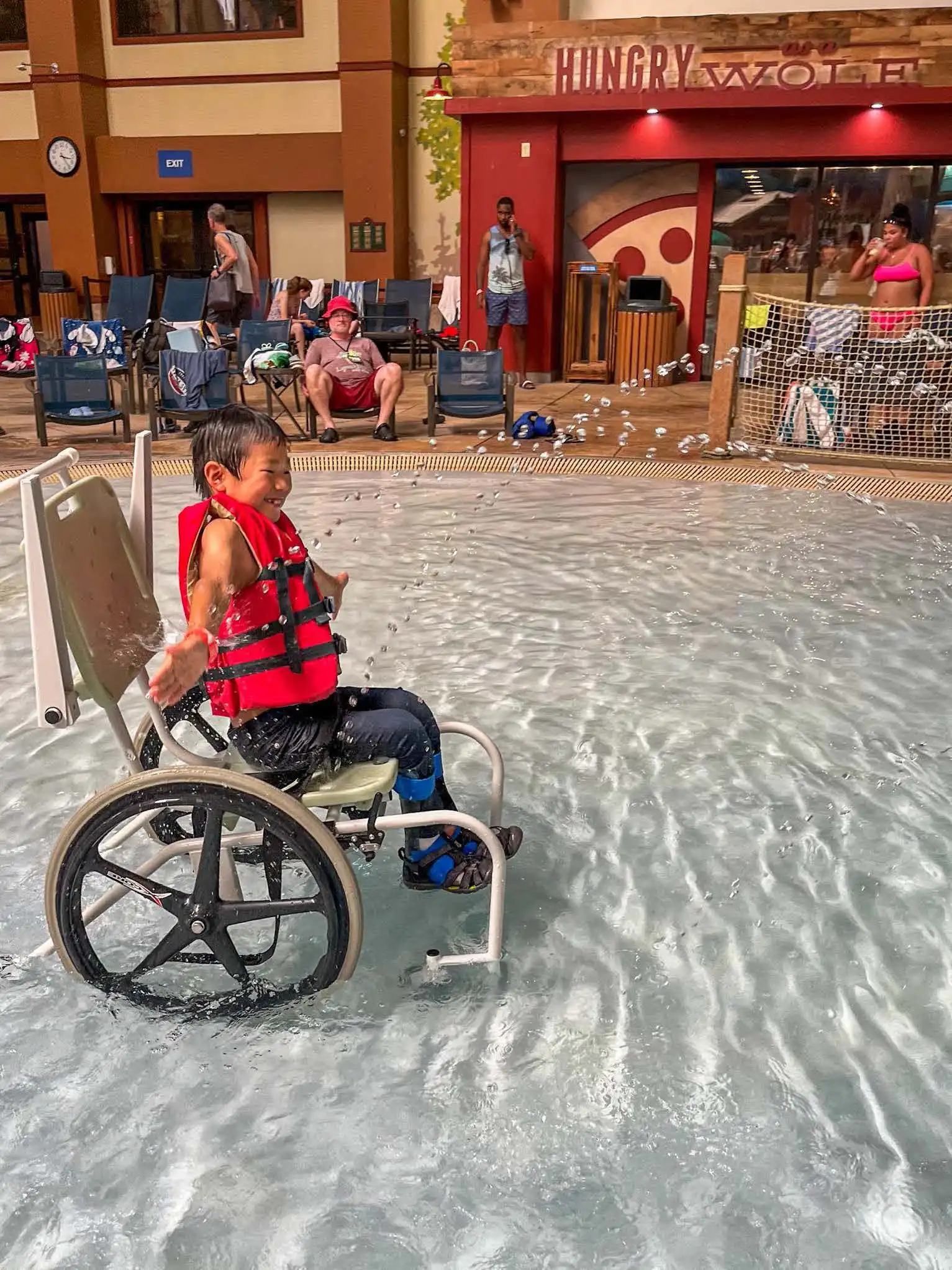 The image shows a child in a wheelchair splashing water in a shallow pool. The child is wearing a red life jacket and smiling with joy, clearly enjoying the water experience. The wheelchair is designed for water use, with large wheels suited for the pool environment. Behind the child, other people are seated near the poolside, with towels and swim gear visible, and a restaurant or food stand in the background labeled "Hungry Wolf." The overall scene is lively and inclusive, highlighting the accessibility of the pool for people with mobility challenges