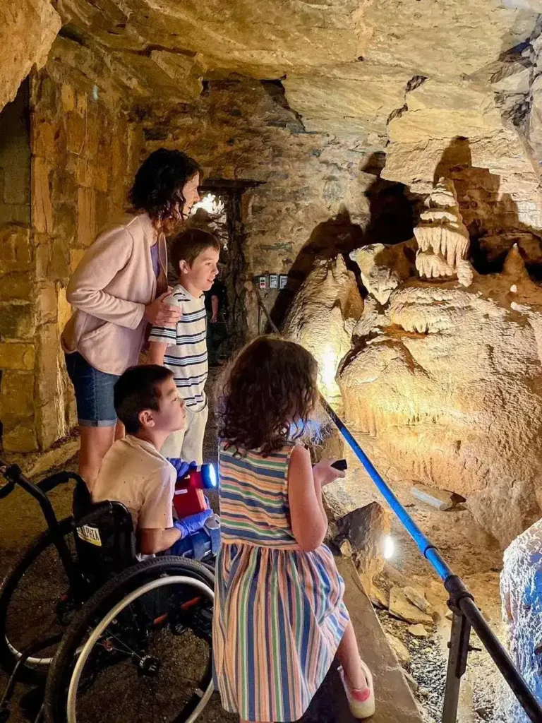 A family is exploring the inside of a cave. The group includes a woman and three children. One of the children is sitting in a wheelchair, and the other two are standing, gazing at the cave's rock formations. The lighting highlights the natural textures of the cave walls, creating a warm and intriguing atmosphere. One of the children is holding a flashlight, adding to the sense of adventure. The cave's rock formations appear to be stalactites and stalagmites, contributing to the mysterious and beautiful surroundings. The scene captures a moment of curiosity and discovery.