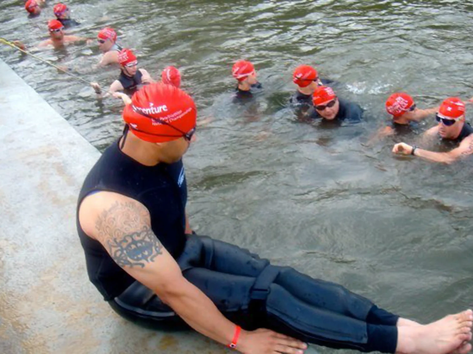 The image shows a group of swimmers preparing to enter the water. They are all wearing red swim caps, black wetsuits, and goggles, indicating participation in a swim event. One swimmer, in the foreground, is sitting on the edge of the platform, ready to enter the water. The swimmer has a visible arm tattoo and is positioned with their legs extended over the water. Several other swimmers are already in the water, holding onto a rope and waiting. The setting appears to be part of a triathlon or open-water swim race.