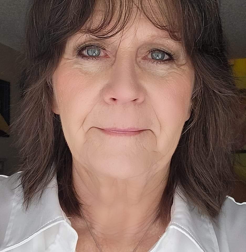 Close-up portrait of a woman with shoulder-length dark hair, light blue eyes, and a calm expression. She is wearing a white collared shirt and looking directly at the camera.