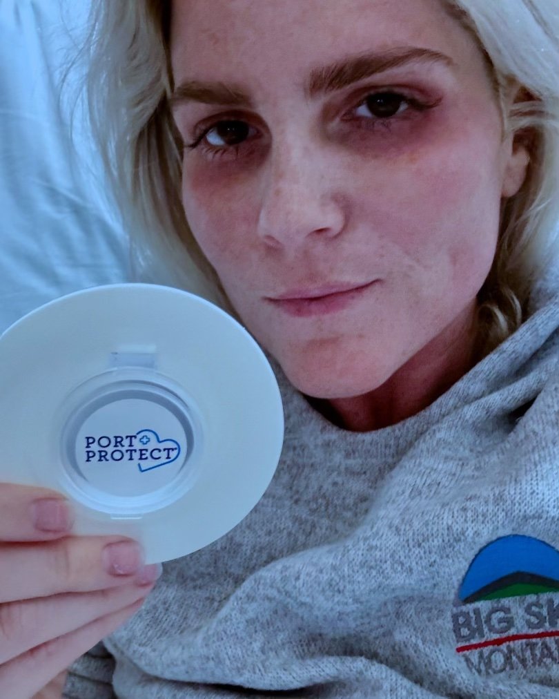 A person with light skin and blonde hair holds a circular medical device labeled "Port Protect" close to their face. They are wearing a gray sweatshirt with a logo reading "Big Sky Montana" on it. The person has a neutral expression and is positioned against a light-colored background.