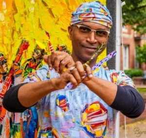 A man in colorful, vibrant clothing stands in front of a bright, textured painting. He is wearing glasses and a matching colorful headscarf. His shirt has abstract patterns with bold reds, yellows, and blues, complementing the painting behind him, which has rich yellow tones. He holds two paintbrushes crossed in front of him, smiling with confidence and creativity. The overall scene is lively and full of color, reflecting an artistic, expressive personality.