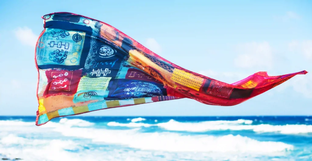 A colorful scarf with abstract, native-inspired designs and vibrant patterns blowing in the wind against a background of a blue sky and ocean waves. The scarf features a mix of blue, red, yellow, and orange patches with intricate symbols, adding a dynamic and cultural feel to the scene