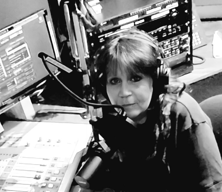 A black-and-white photograph of a woman in a radio studio. She is wearing headphones and is positioned in front of a microphone. Multiple screens and audio equipment surround her, indicating a professional broadcasting setup. The woman is looking directly at the camera with a focused expression. The image captures a moment of concentration, likely during a live or recorded radio session.