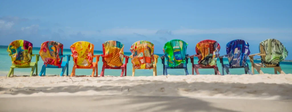 A row of colorful beach chairs draped with vibrant, patterned silk scarves on a sandy beach, facing a calm turquoise sea under a clear blue sky. Each scarf features unique, abstract designs and bright colors, reflecting Elisa Lejuez’s signature style.