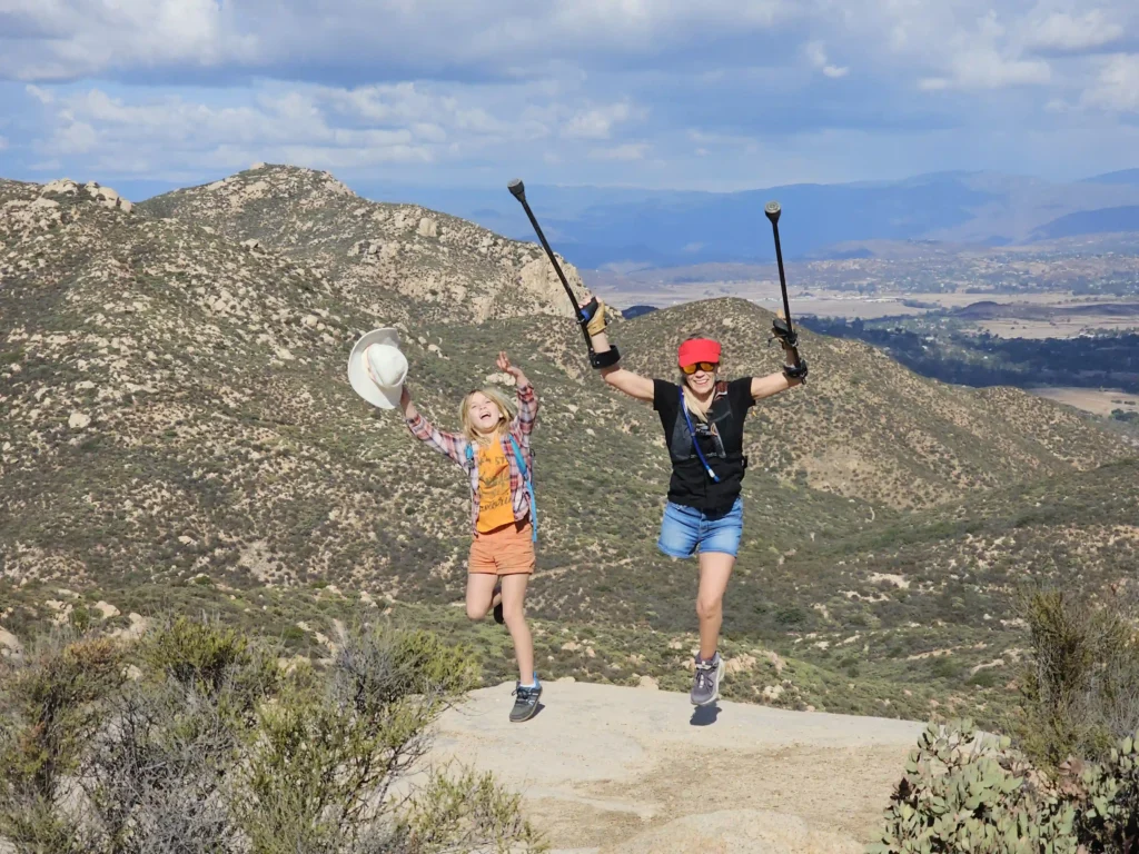 Image of a joyful hiking moment, with a woman using crutches and a child leaping together on a scenic mountain trail under a blue sky.