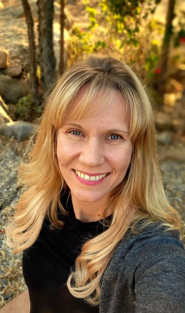 Portrait of a smiling woman in a sunny outdoor setting, with a background of trees and warm natural light.