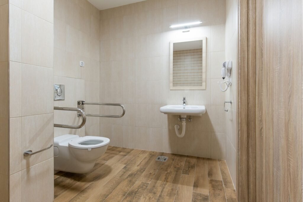 An accessible bathroom featuring a wall-mounted toilet with grab bars on both sides, a wall-mounted sink, and wooden-style flooring with beige tiled walls, designed for wheelchair users or individuals with mobility challenges.