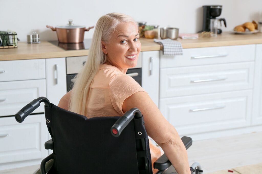 A smiling woman with long blonde hair seated in a wheelchair in a modern kitchen with white cabinetry and light wooden countertops, emphasizing inclusivity and accessibility in home design.