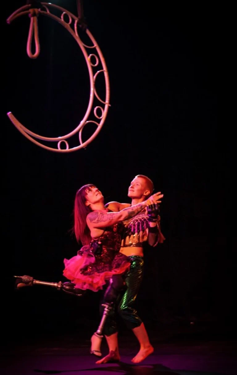 Two performers gracefully dance under a crescent-shaped aerial hoop, one wearing a pink and black tutu and the other in shimmering green pants, showcasing a prosthetic leg and creative costuming in a dynamic and empowering performance