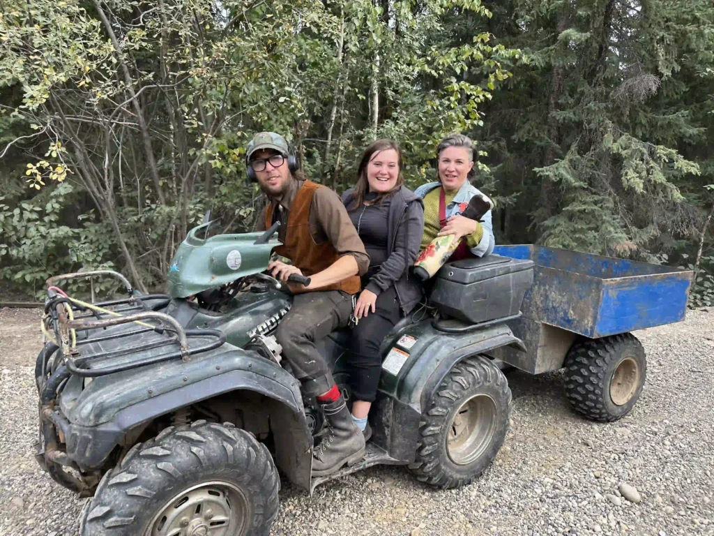 Three people sitting on an all-terrain vehicle (ATV) with a blue trailer attached, surrounded by a forested area. The person in front is operating the ATV, while the two individuals behind are smiling and enjoying the ride